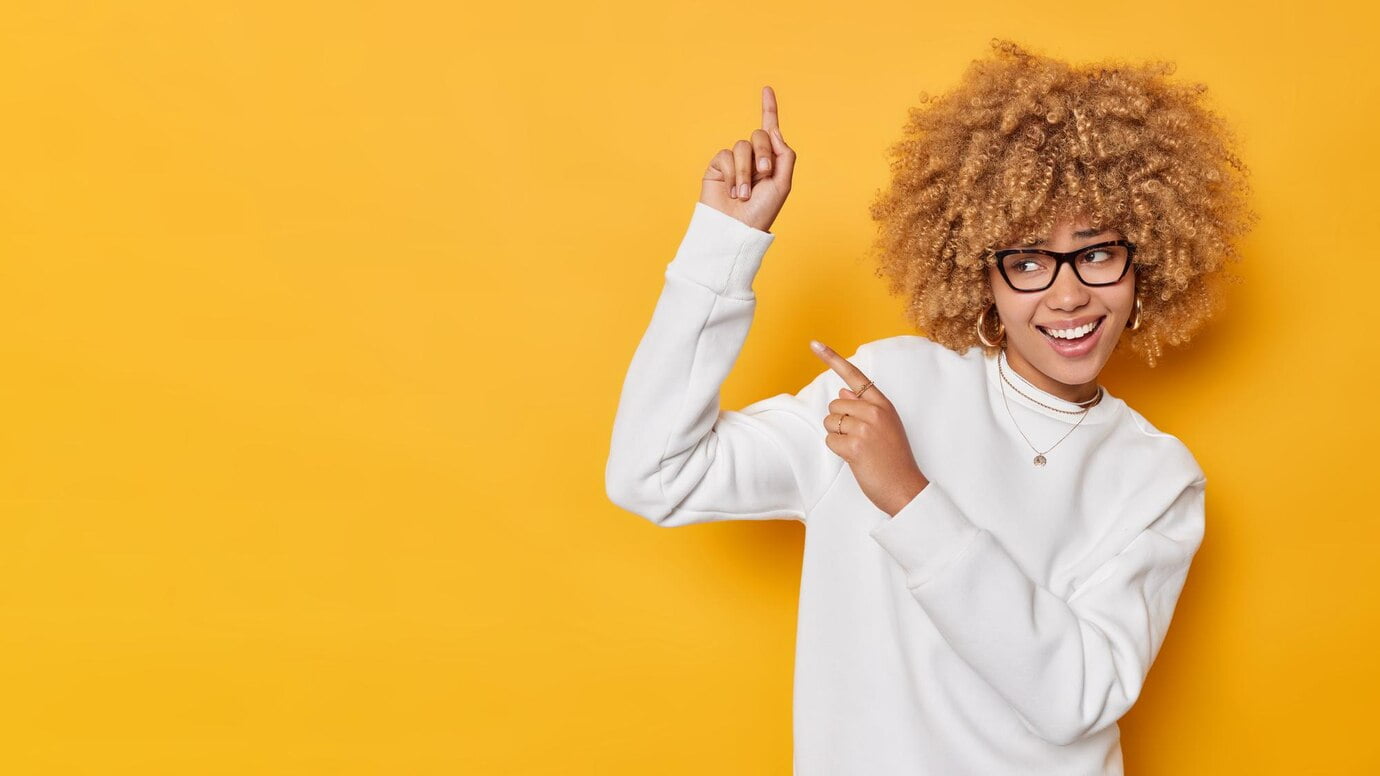 cheerful curly haired adorable woman points against blank space shows information banner demonstrates advertisement wears spectacles white casual jumper isolated yellow background 273609 60809